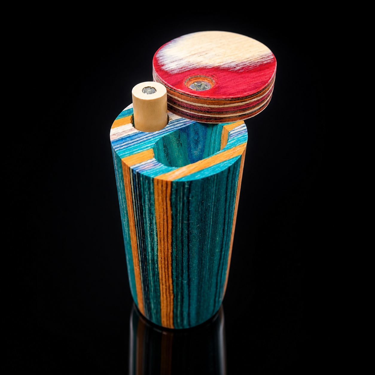 A colorful wooden cylindrical container with a lid, photographed on a black reflective surface. The container features vertical wood grain stripes in teal, blue, orange, and brown, with a small beige cylindrical insert.