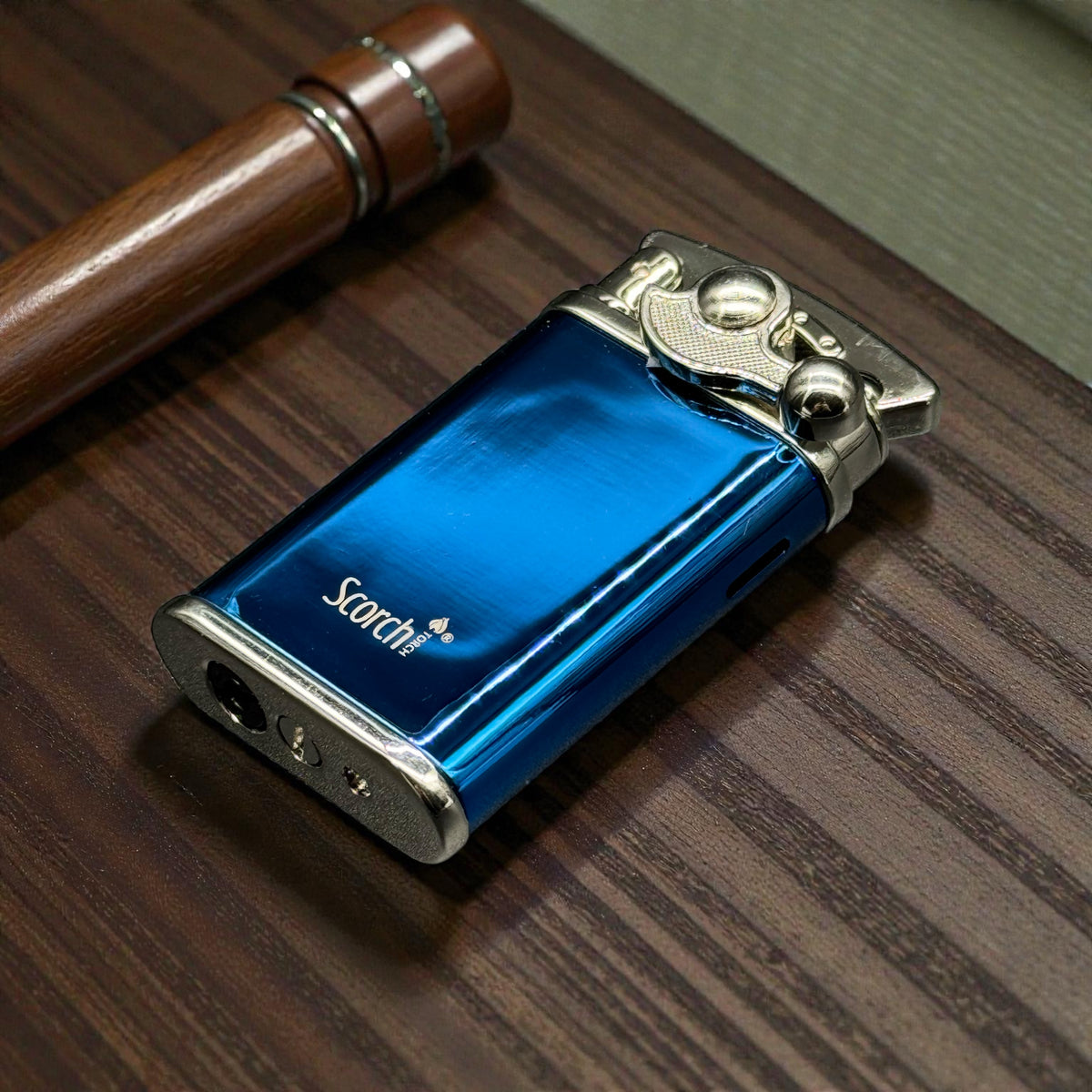 A metallic blue lighter with silver accents and the brand name 'Scotch' printed on the side, resting on a dark wood-grained tabletop next to a wooden pen.