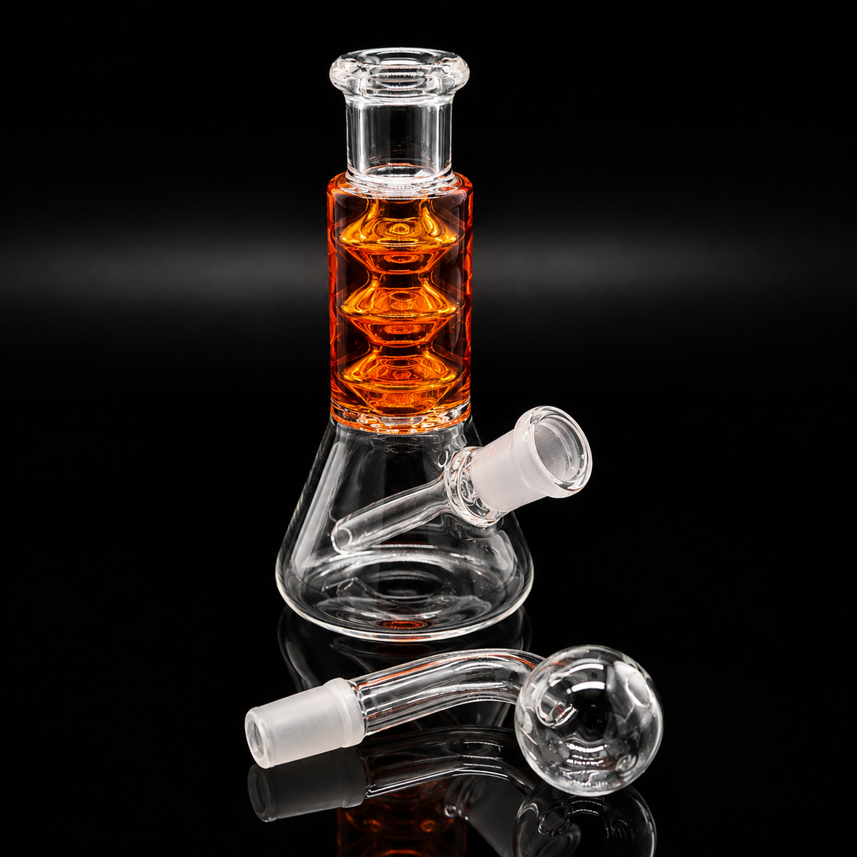 Clear glass beaker-style water pipe with an amber-orange liquid-filled chamber and a clear glass neck, shown on a black reflective background with two detachable glass mouthpieces placed in front.