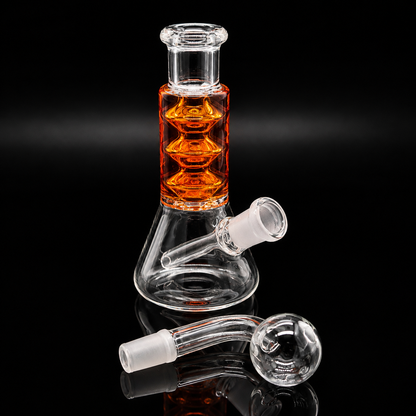 Clear glass beaker-style water pipe with an amber-orange liquid-filled chamber and a clear glass neck, shown on a black reflective background with two detachable glass mouthpieces placed in front.