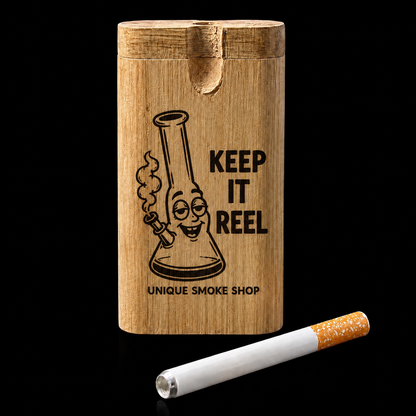 A rectangular wooden cigarette case with a lid, featuring a black engraved cartoon bong and the text 'KEEP IT REEL' and 'UNIQUE SMOKE SHOP'. A white cigarette with an orange tip lies in front on a black background.