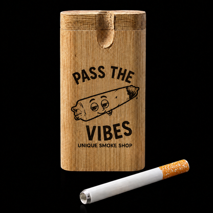 A cylindrical wooden smoking accessory holder with a carved slot lid, engraved with the text 'PASS THE VIBES UNIQUE SMOKE SHOP' and a cartoon joint graphic, shown on a black background with a cigarette resting in front.