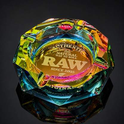 A faceted iridescent glass or acrylic smoking ashtray with a gold-toned engraved center reading 'The Natural Way to Roll RAW Since 2005,' shown on a dark reflective surface against a black background.