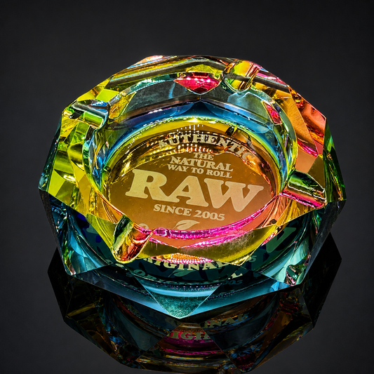 A faceted iridescent glass or acrylic smoking ashtray with a gold-toned engraved center reading 'The Natural Way to Roll RAW Since 2005,' shown on a dark reflective surface against a black background.