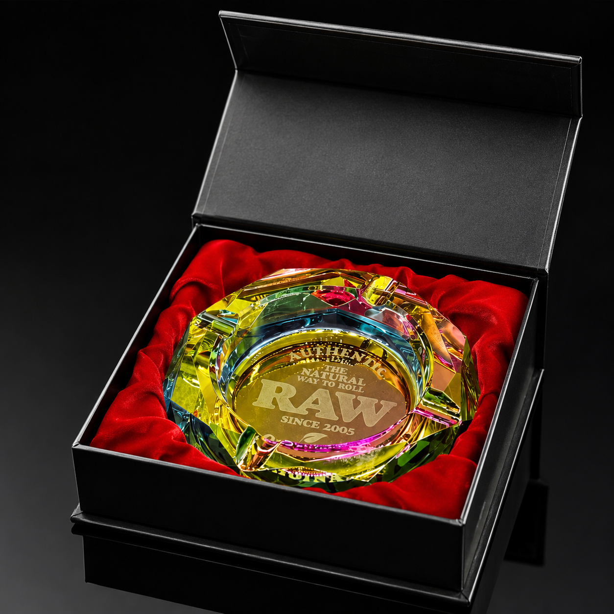 A clear acrylic and glass ashtray with colorful iridescent rainbow reflections, engraved with 'RAW SINGLES CLUB' and 'AUTHENTIC RAW', presented in an open black gift box with red velvet lining.