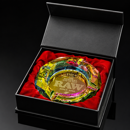 A clear acrylic and glass ashtray with colorful iridescent rainbow reflections, engraved with 'RAW SINGLES CLUB' and 'AUTHENTIC RAW', presented in an open black gift box with red velvet lining.