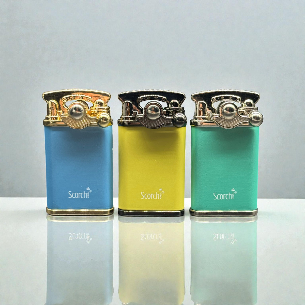 Three portable pocket lighters in blue, yellow, and mint green, each with a polished gold-toned metal flame tip and the 'Scorchi' brand logo printed on the front, displayed on a reflective white surface against a light gray background.