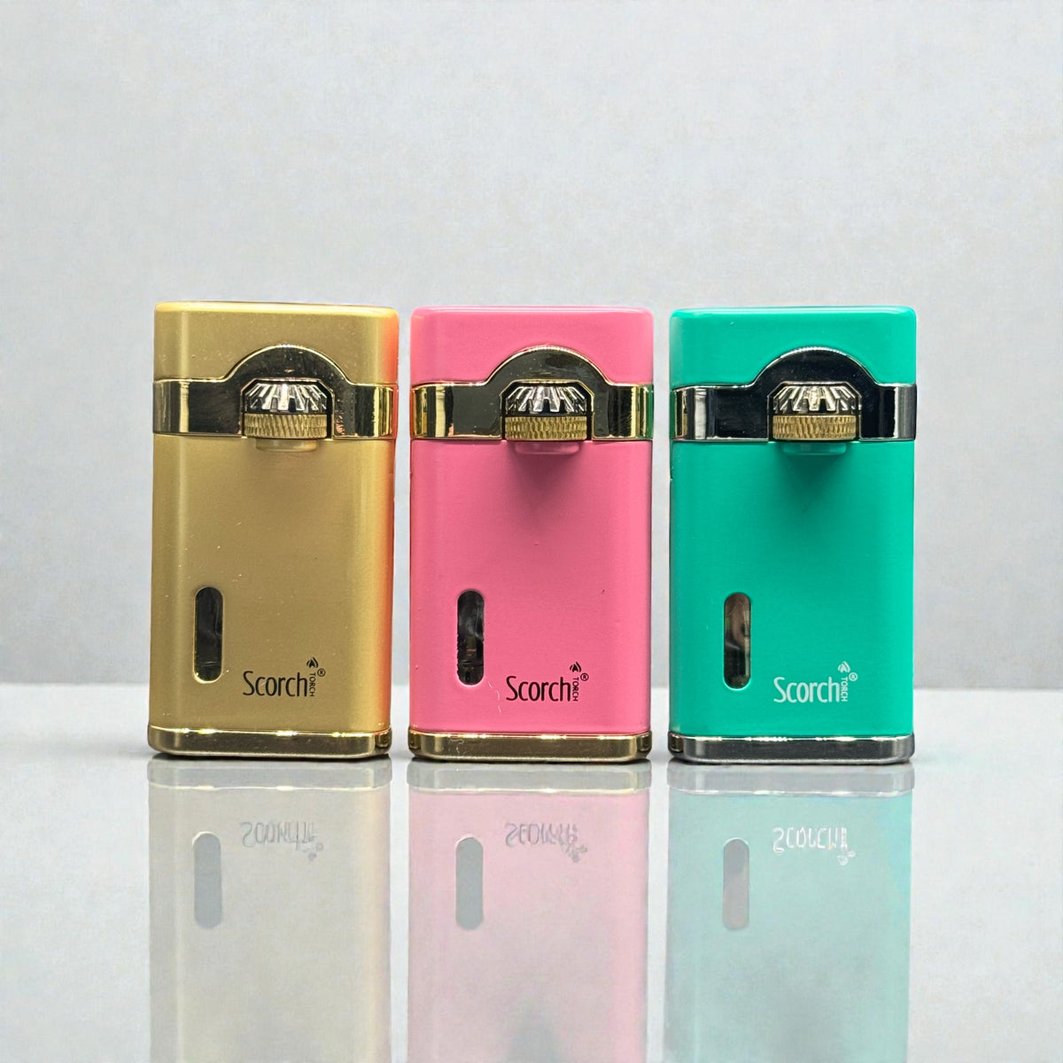 Three portable electronic lighters in gold, pink, and teal green, lined up side-by-side on a reflective white surface against a light gray background.
