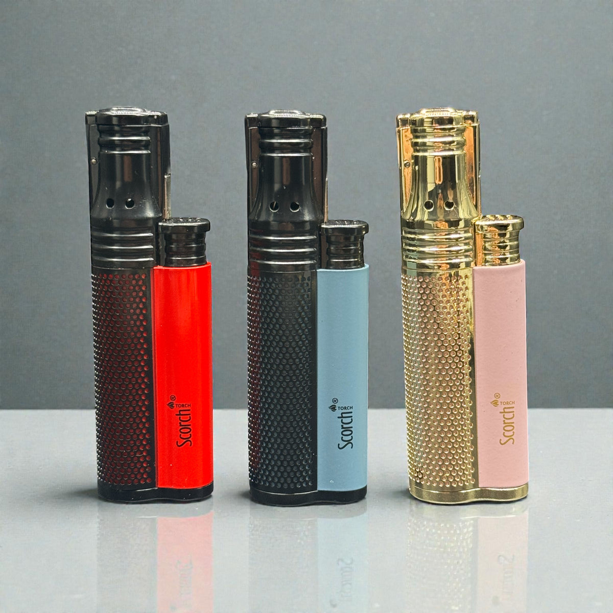 Three Sorthi pocket lighter replicas displayed side-by-side on a reflective gray surface against a dark gray background. The set includes a dark brown and red lighter, a black and light blue lighter, and a gold and pink lighter.