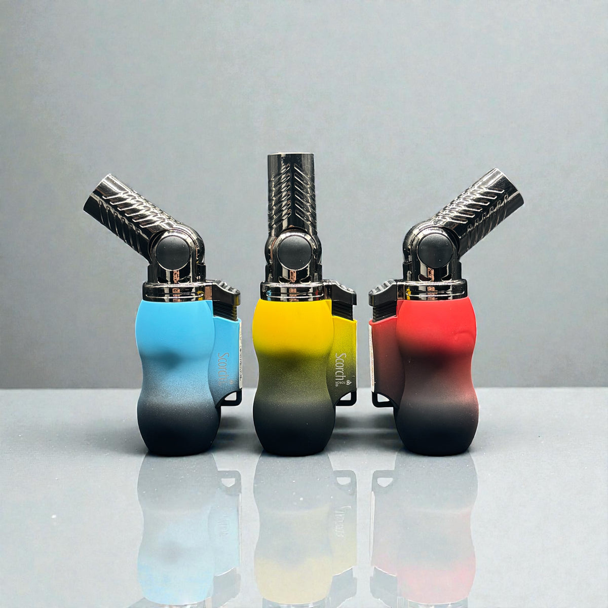 Three portable flame torch lighters in blue, yellow, and red gradient finishes, arranged side-by-side on a reflective gray surface against a neutral gray background.