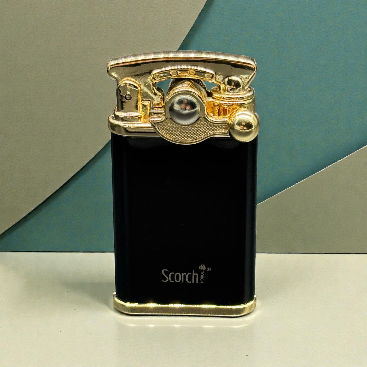 A black Scorch portable lighter with a polished gold-toned metal top and base, standing upright on a light surface against a geometric teal and gray background.