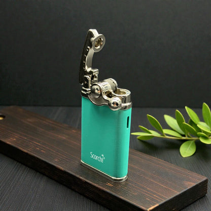 A turquoise metal lighter with a silver flip-up flame guard, standing on a dark wooden block with green leaves in the background.