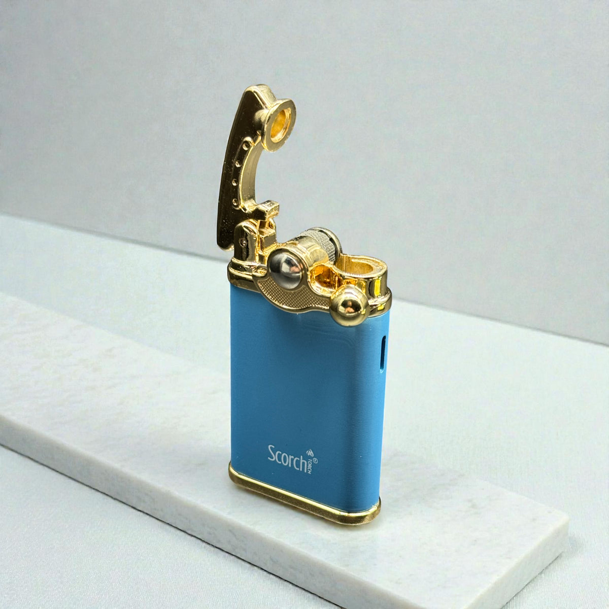 A blue and gold portable electric lighter with the lid open, resting on a white marble slab against a light gray background. The brand name 'Scorch' is printed on the front.