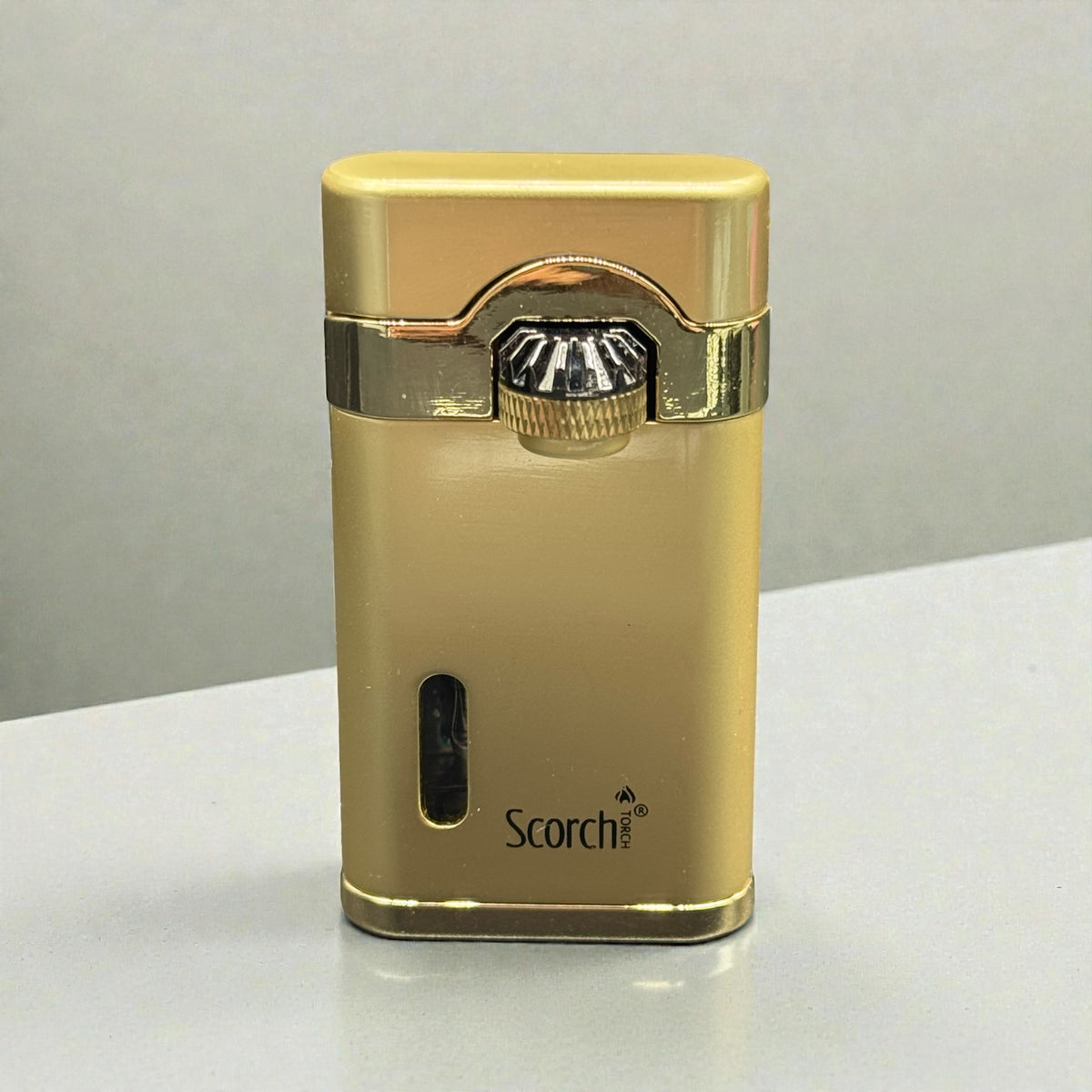 A gold metallic Scorch brand lighter with a polished chrome ignition button, standing upright on a light gray surface.