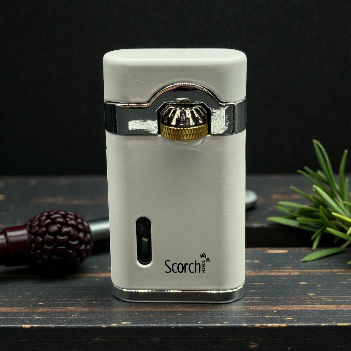 A white Scorch electronic cigar torch lighter with a silver metal band and a gold-colored ignition knob, displayed on a dark wooden surface with a dark background and decorative props.