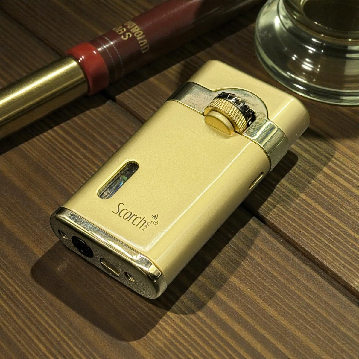A cream-colored Scorch electric lighter with polished silver and gold accents, resting on a dark wood tabletop next to a red and gold pen and a glass.