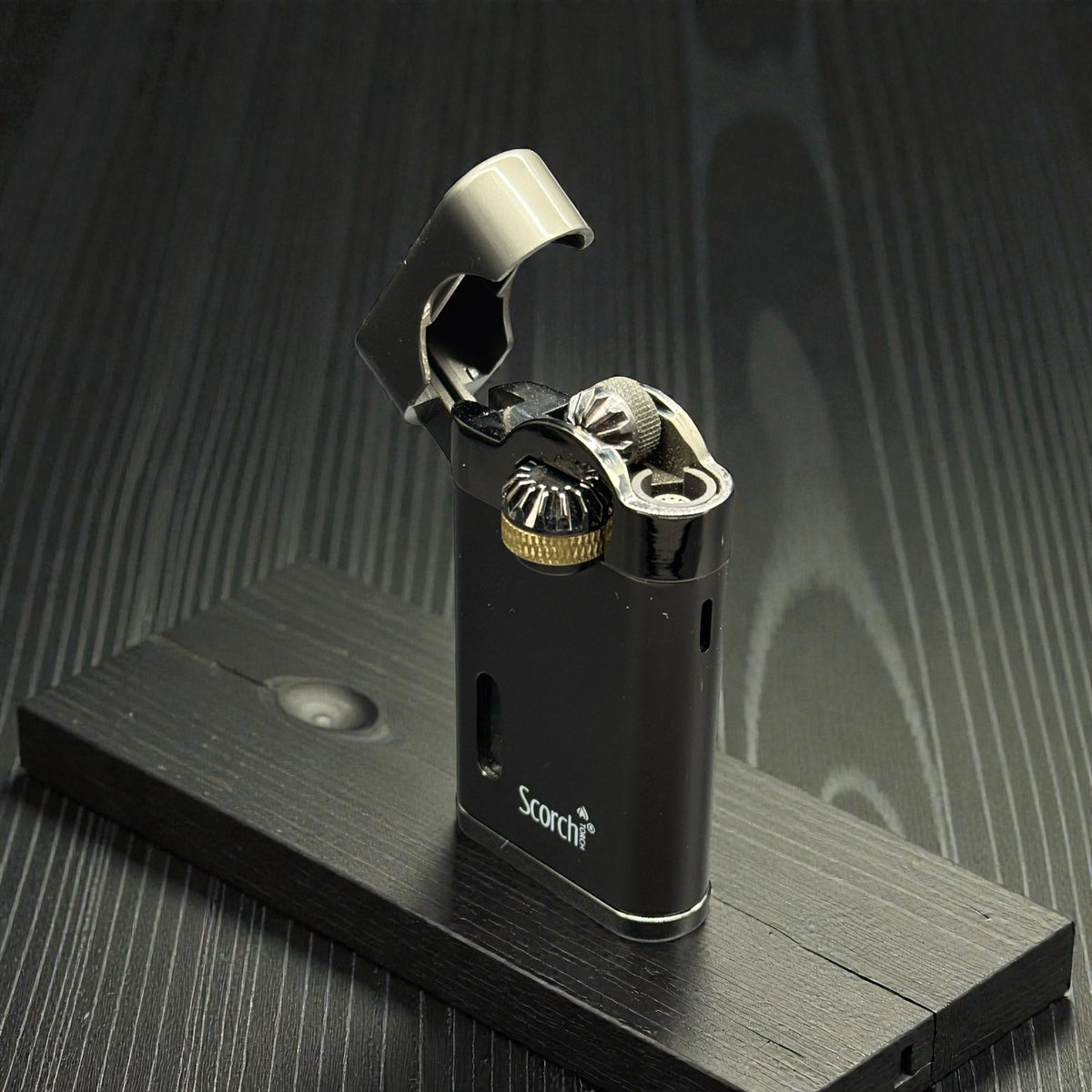 A black metallic pocket lighter with a silver flip-up lid and gold-toned ignition detail, standing on a dark wooden display block against a dark wood-grain background. The brand name 'Scotch' is printed on the lower front.