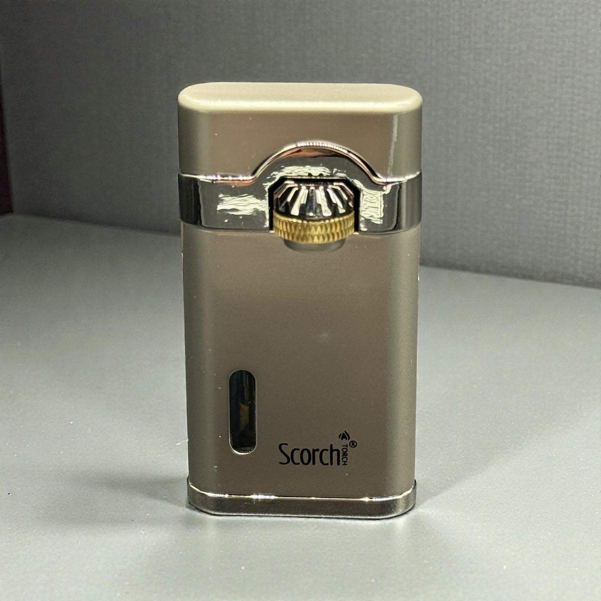 A metallic silver and gold Scorch brand lighter standing upright on a gray surface against a gray background.