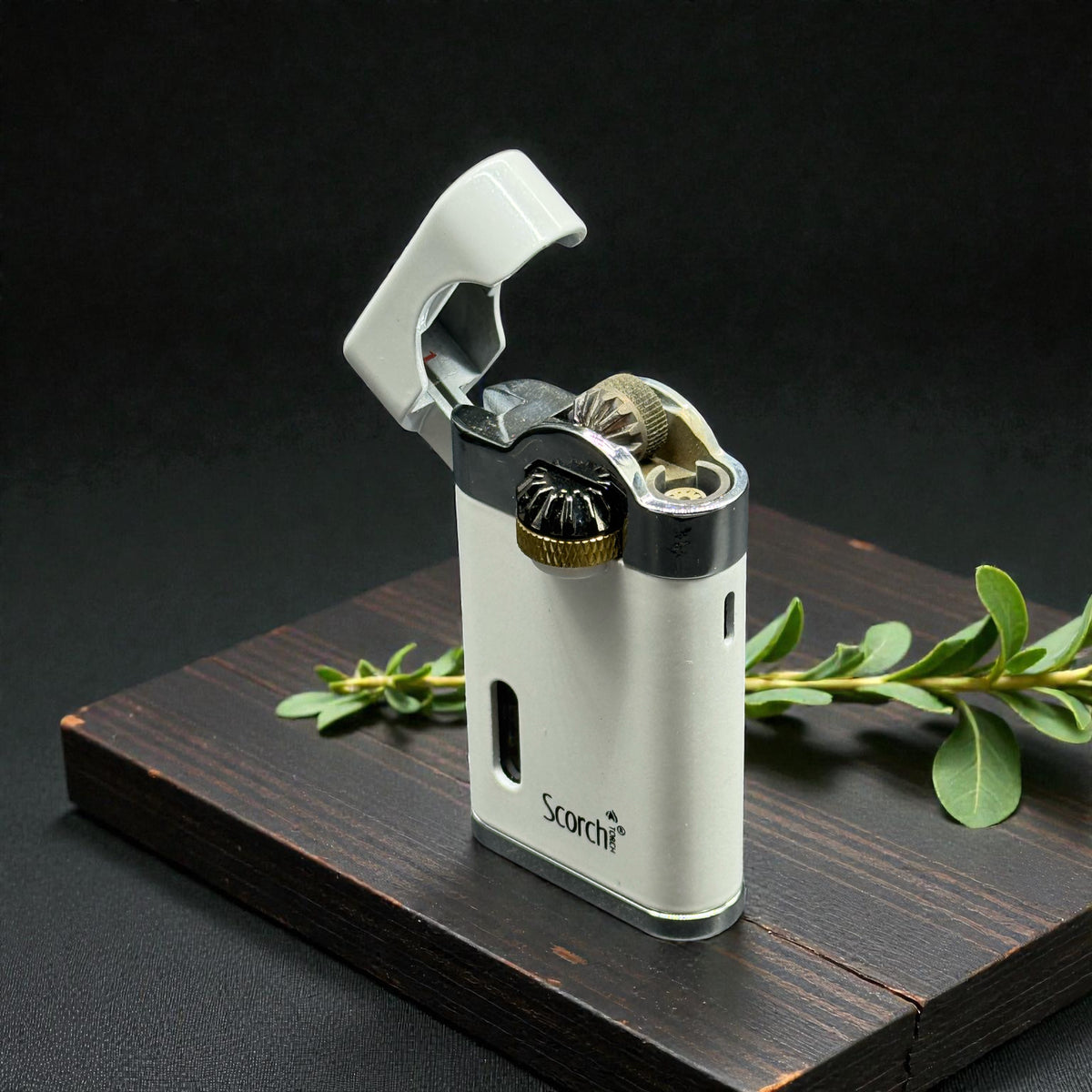 A white and silver Scarf brand lighter with a gold ignition button, displayed on a dark wooden block with green leaves against a black background.