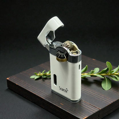 A white and silver Scarf brand lighter with a gold ignition button, displayed on a dark wooden block with green leaves against a black background.