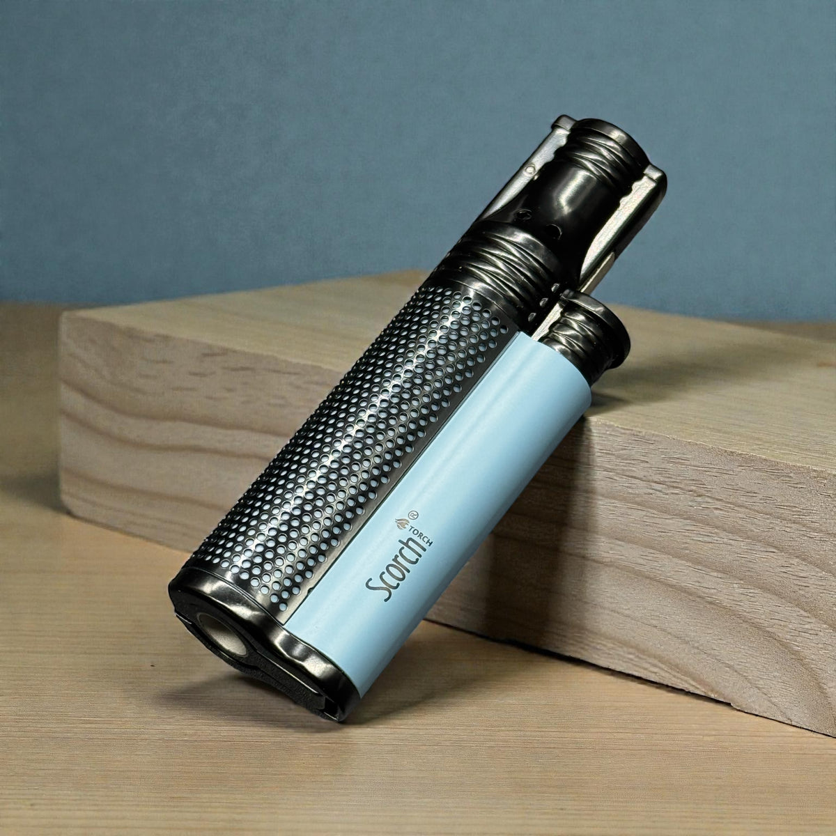 A light blue and black carbon-fiber-textured lighter branded 'Scorch' resting on a wooden block against a blue-gray background.