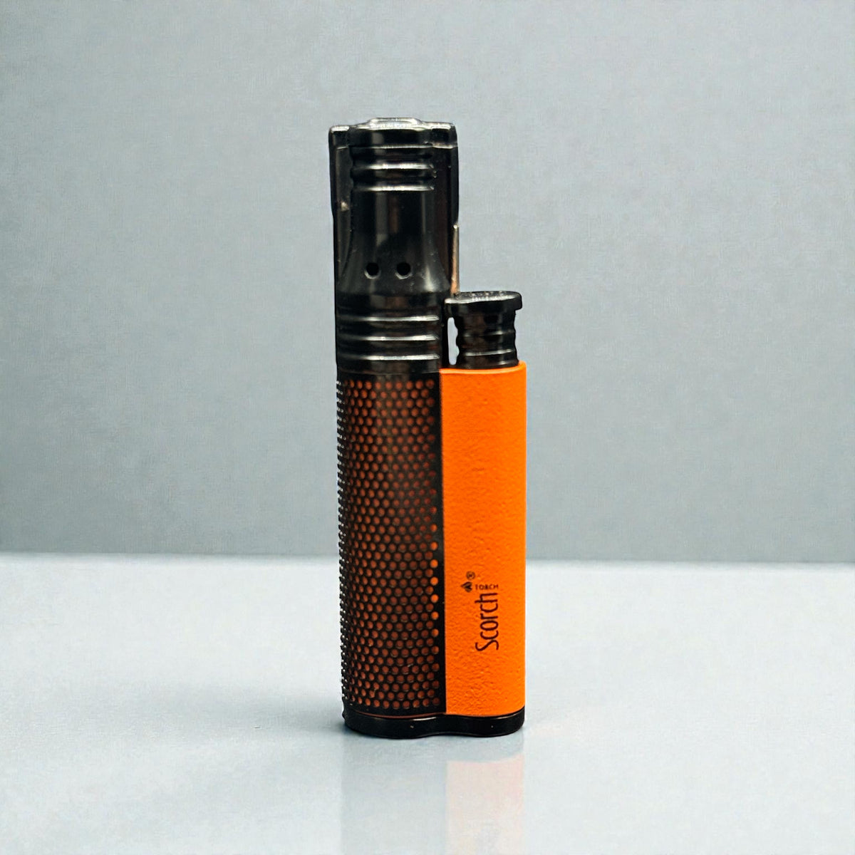 A dual-color lighter standing upright on a light gray reflective surface against a gray background. The lighter features a textured dark brown metallic body on the left and a bright orange metallic body on the right, with the brand name 'Sorthi' printed vertically in black on the orange section.