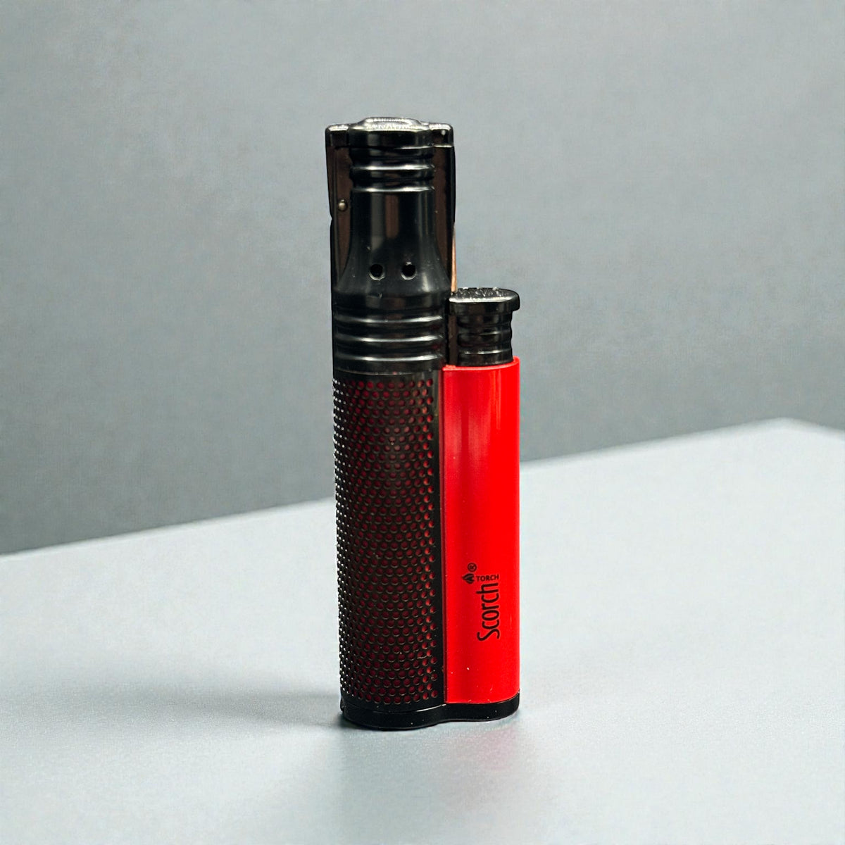 Two Satchi brand pocket lighters standing upright on a light gray surface against a gray background. One lighter is black with a dark textured grip, and the other is bright red with a black top and bottom.