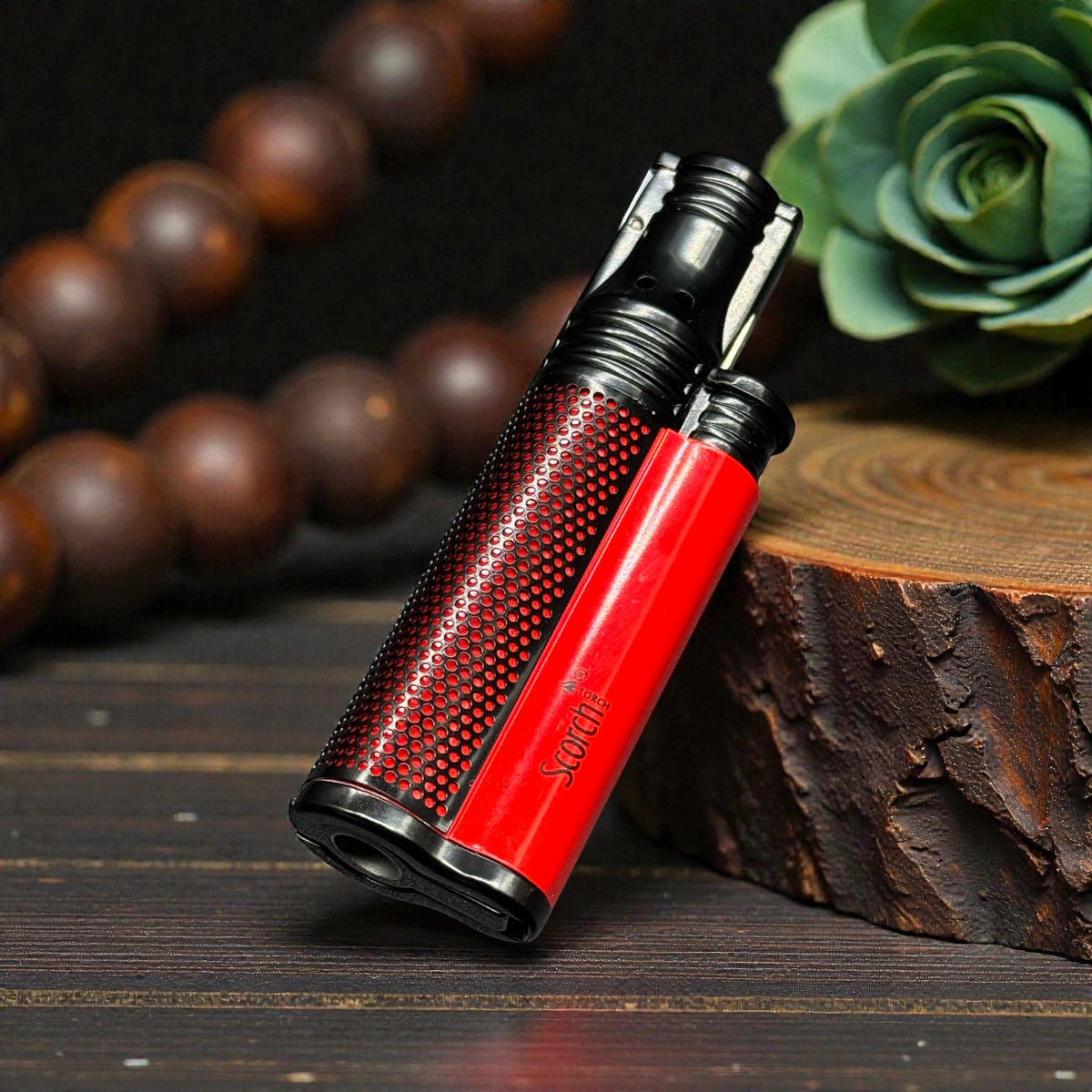 A red and black textured lighter with the brand name 'Socra' printed on the side, resting on a rustic wooden block with a dark wood background and blurred decorative beads and a succulent.