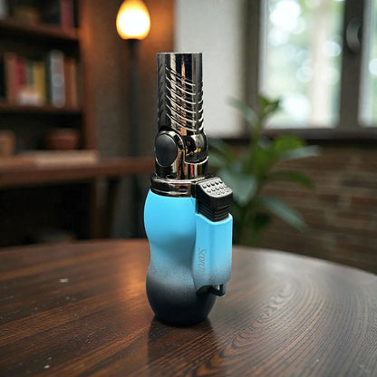 A blue and black torch lighter with a metallic nozzle stands on a dark wooden table in a softly blurred indoor setting with a bookshelf and window in the background.