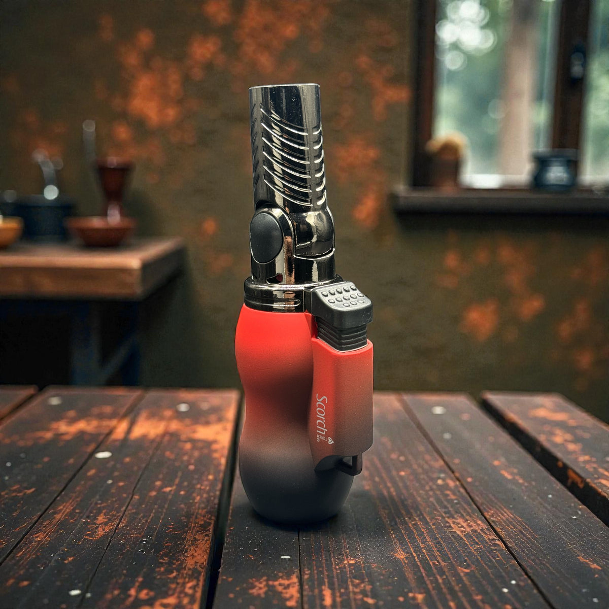 A modern pocket torch lighter with a red-to-black gradient body and a polished chrome nozzle, standing on a rustic wooden table in a warm, atmospheric setting.