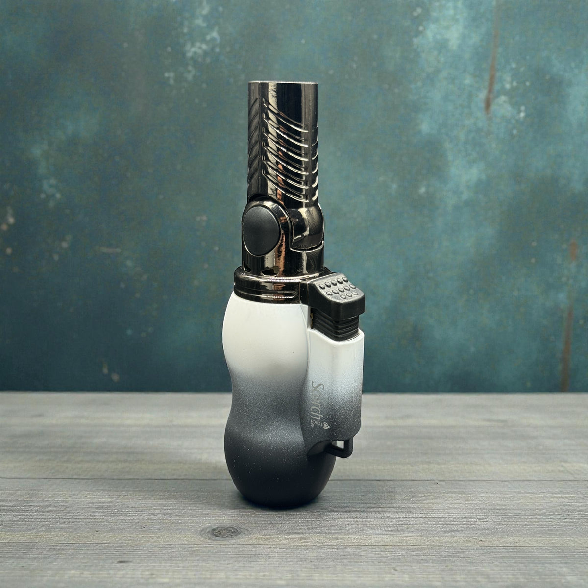 A metallic torch lighter with a black-to-white gradient handle, standing upright on a gray wooden surface against a textured teal-blue background.