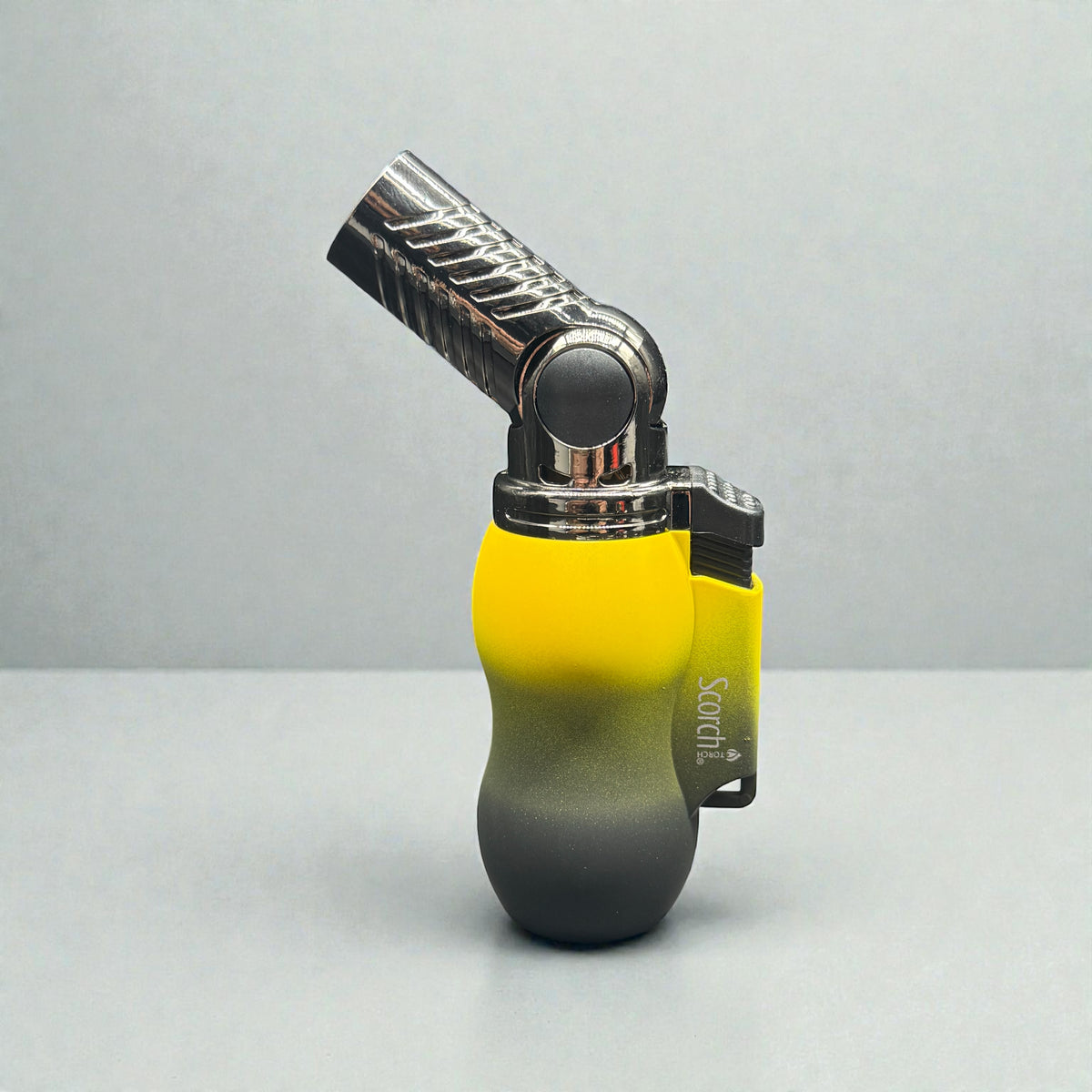 A portable torch lighter with a gradient body transitioning from bright yellow at the top to dark gray/black at the bottom, featuring a metallic silver and black swivel torch head. The lighter is standing upright on a light gray surface against a matching gray background.