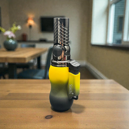 A handheld torch lighter with a yellow-to-black gradient body and a chrome atomizer tip, standing on a wooden table in a blurred indoor room.