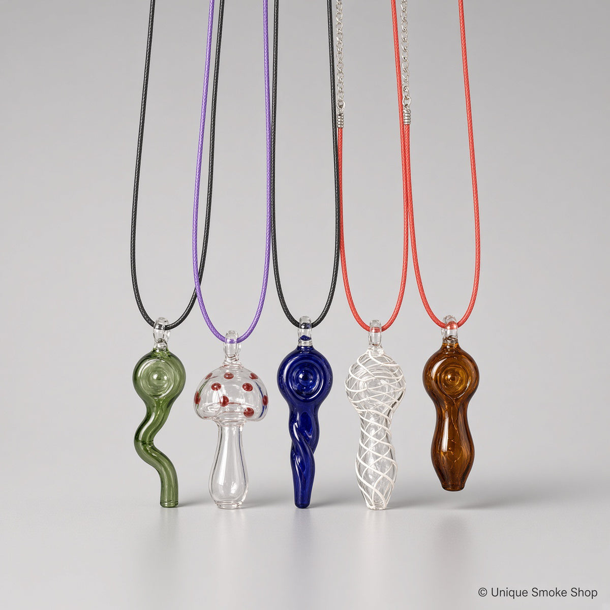Assorted mini hand-blown glass pipe necklaces in different colors and designs including green wavy, mushroom, blue spiral, clear swirl, and amber styles hanging on cords