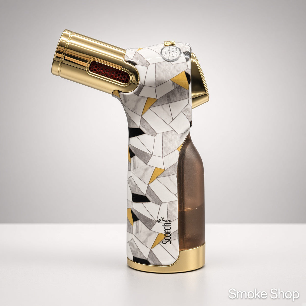 A Stocchi torch lighter with a geometric mosaic pattern in white, gray, black, and gold, featuring a translucent brown fuel tank and gold accents, set against a neutral gray background.