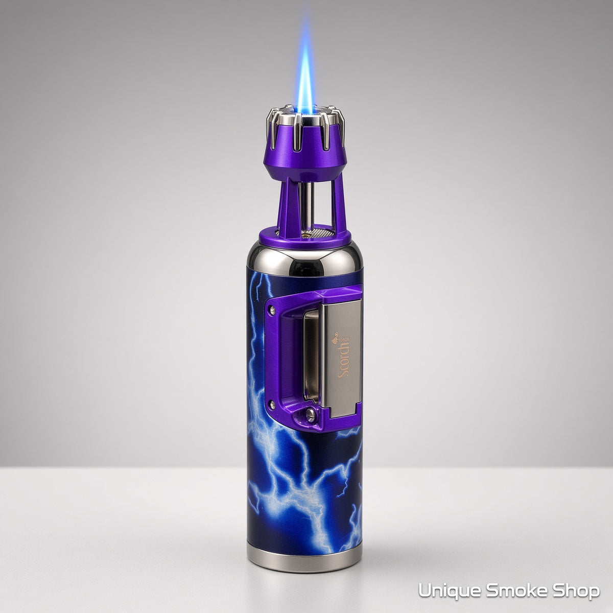 A metallic purple and blue lightning-pattern lighter with a visible blue flame at the nozzle, photographed on a clean light gray studio background. The lighter has a chrome-toned base and a purple side handle.